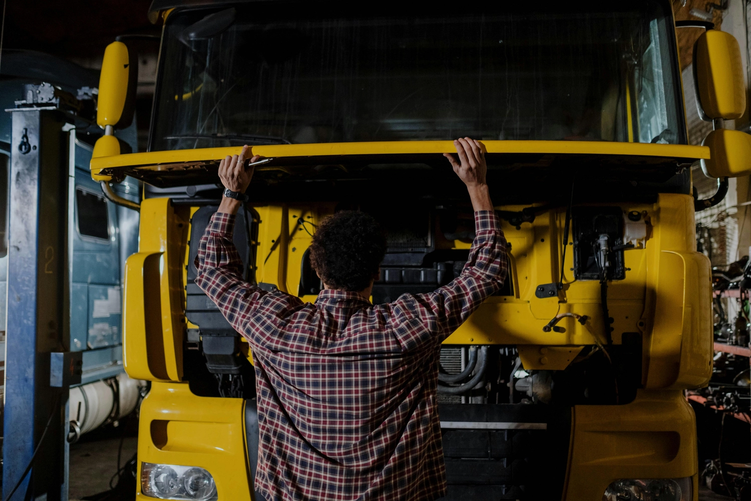 Mobiler Gasservice und Serviceleistung – Serviceleistungen rund um Wohnmobil und Wohnwagen – Mann im karierten Hemd öffnet die Motorhaube eines gelben Lastwagens in einer schwach beleuchteten Garage