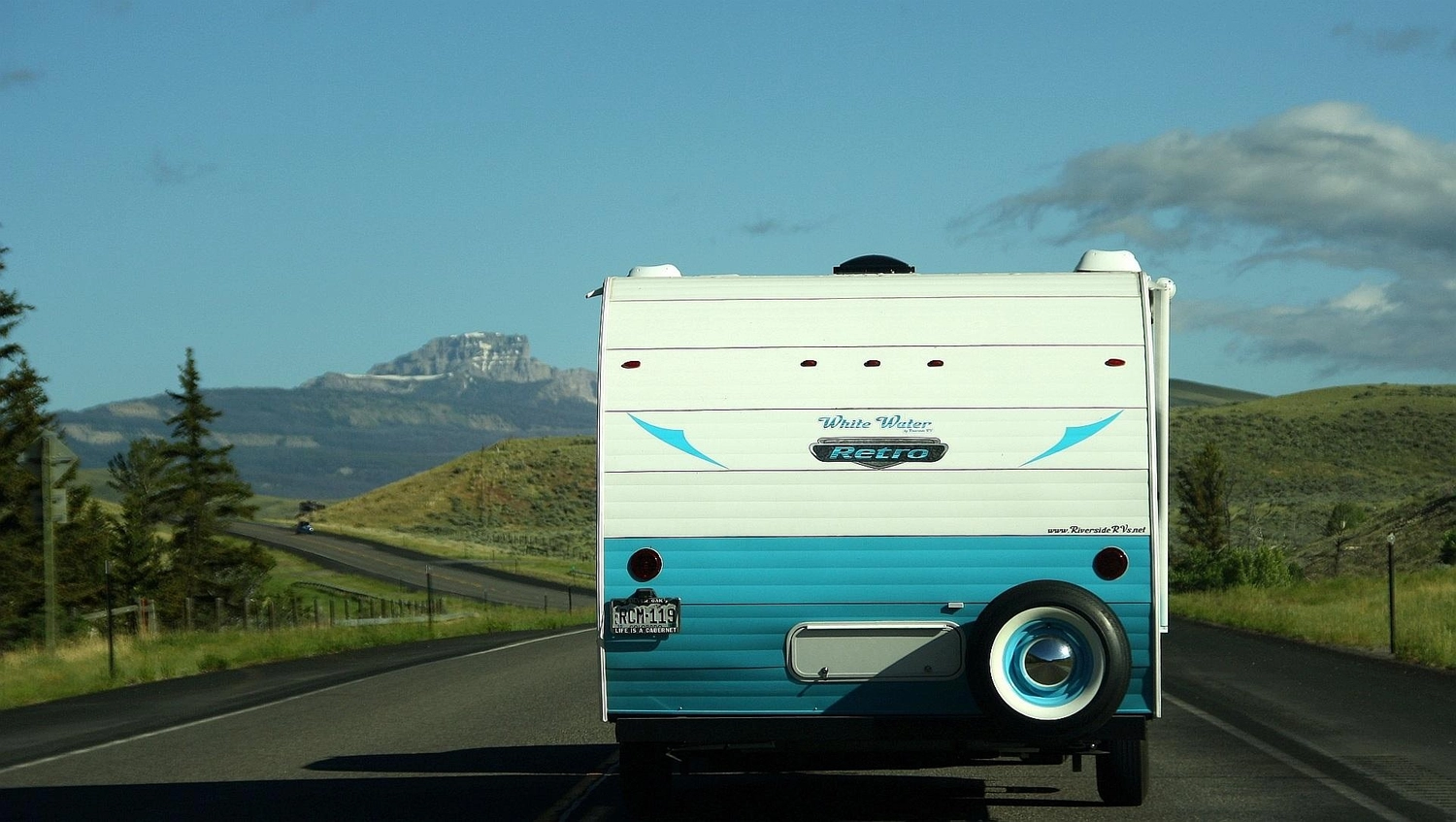 Mobile Gas Service und Serviceleistungen - serviceleistungen rund um Wohnmobil und Wohnwagen – Rückansicht eines blau-weißen Retro-Wohnmobils, das auf einer Autobahn fährt, mit Bergen und Bäumen im Hintergrund